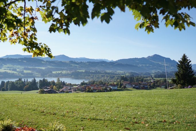 Landschaft mit grüner Wiese, Blick auf Berge und ein Dorf am See.